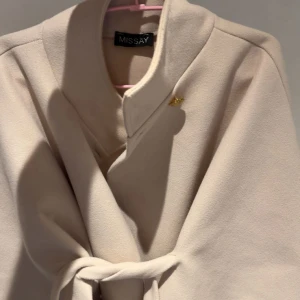Beige kappa med bälte från Missay - Stilren beige kappa från Missay med knytbälte i midjan och guldknapp vid kragen. Jackan har en elegant, lös passform och hög krage. Perfekt för dig som vill ha en klassisk look med modern twist.