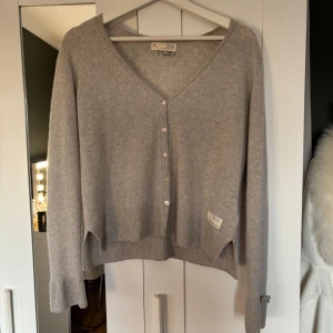 Odd Molly kofta - Superfin ljusgrå kofta med v-ringning och knappar framtill. Baksidan har ett diskret stickat mönster och siffran 657 broderad upptill. Ärmarna har bred mudd och en söt liten rosett. Perfekt för lager-på-lager och mysiga dagar. Storlek 2 i Odd Molly storlekar vilket motsvarar M❤️