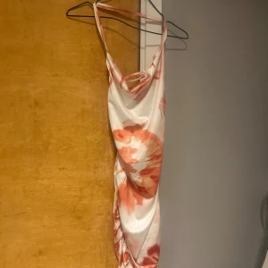 Vit och rosa långklänning med mönster - Säljer en snygg långklänning i vitt med rosa mönster. Klänningen har smala axelband och en draperad halsringning. Perfekt för sommarens alla tillfällen. Materialet känns mjukt och följsamt.