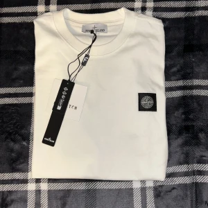 Vit t-shirt från Stone Island - Vit t-shirt från Stone Island med klassisk rund hals och ikonisk logotyp-patch på bröstet. Tillverkad i mjuk bomull för en skön känsla. Perfekt för dig som gillar stilrena och exklusiva plagg.