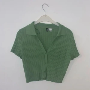 Grön ribbad croppad blus med krage - Säljer en ljusgrön ribbad croppad blus med kort ärm och klassisk krage. Blusen har knappar framtill och är i ett mjukt, stretchigt material. Perfekt för vår och sommar!