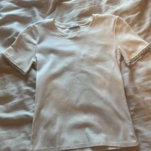 Vit t-shirt med pärldetaljer från Zara - Säljer en stilren vit t-shirt från Zara med korta ärmar och snygga pärldetaljer längst ut på ärmsluten. Perfekt för dig som vill ha en enkel men ändå unik look. Materialet känns mjukt och skönt mot huden.