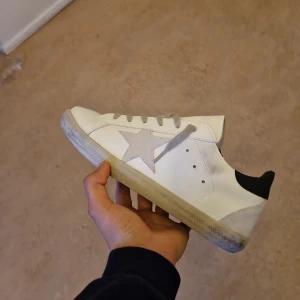 Golden goose  - Säljer ett par vita sneakers från Golden Goose med grå stjärna på sidan, grå snörning och svart hälparti. Skorna har en klassisk låg profil och platt sula. Perfekta för dig som gillar en stilren men ändå unik look.