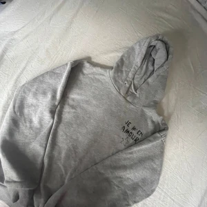 Grå hoodie från Gina Tricot med tryck - Säljer en grå hoodie från Gina Tricot med svart texttryck 'JE M'EN AMOUR' och en stjärna på bröstet och ryggen. Tröjan har huva och långa ärmar, perfekt för en avslappnad stil men funkar också till en styling. Säljer då jag har två likadana. 