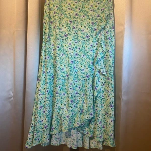 Blommig omlottkjol från Lindex - Säljer en blommig omlottkjol från Lindex i grönt, lila och gult. Kjolen har volangdetalj längs kanten och är i ett lätt, somrigt material. Perfekt för dig som gillar färg och mönster!