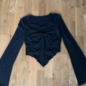 Svart långärmad topp med asymmetrisk nederkant - Trendig svart topp med långa ärmar och unik asymmetrisk nederkant. Toppen har en figurnära passform och en lätt rynkad detalj framtill. Storlek s