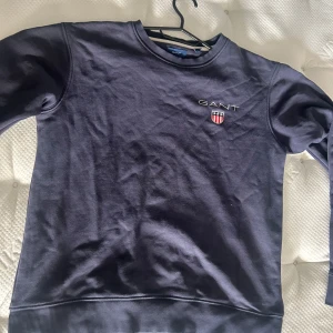 Mörkblå sweatshirt från GANT - Säljer en klassisk mörkblå sweatshirt från GANT med broderad logga och sköld på bröstet. Tröjan har rund halsringning och ribbade muddar vid ärmslut och nederkant. Perfekt för en stilren och avslappnad look.