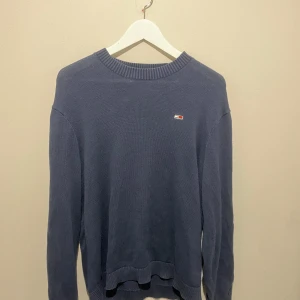 Blå stickad tröja från Tommy Jeans - Mörkblå stickad tröja från Tommy Jeans med rund halsringning och logga på bröstet. Snygga vita band med TOMMY JEANS-tryck längs ärmarna. Storlek är XS men sitter mer som S,M