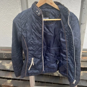Ridjacka från Jackson  - Snygg mörkblå quiltad jacka från Jacson med ståkrage och dragkedja framtill. Jackan har två fickor med dragkedja och ljusa kontrastdetaljer längs kanterna. Perfekt för dig som gillar stilren design och vill ha något som sticker ut lite.