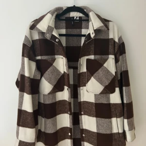 Brun och vit rutig overshirt från H&M Divided - Säljer en brun och vit rutig overshirt från H&M Divided. Jackan har stora bröstfickor med lock, knappar framtill och klassisk krage. Perfekt att slänga över en hoodie eller t-shirt för en avslappnad look.