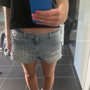 Lågmidjade jeansshorts  - Säljer ett par ljusblå jeansshorts med coola nitar på ena sidan och slitna detaljer på andra. Klassisk femficksmodell med bälteshällor och knappgylf. Perfekta för sommaren och ger en edgy look. Köpta secondhand men med prislappen kvar på, den är dock borttagen men har inte blivit använda! ❗️säljer endast vid bra pris❗️står strl 38 men sitter bra på mig med 34!
