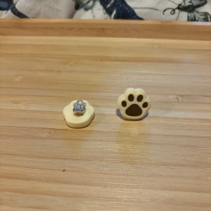 Clips Örhängen med tassmotiv - Säljer ett par söta örhängen formade som djurtassar i vitt med bruna detaljer. Perfekta för dig som älskar djur och vill ha något unikt och lekfullt i din stil.