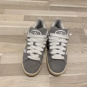 Grå Adidas sneakers med vita detaljer - Säljer ett par grå Adidas sneakers med vita ränder och breda vita skosnören. Skorna har klassisk Adidas-logga på plösen och är låga i modellen. Perfekta för en avslappnad stil.