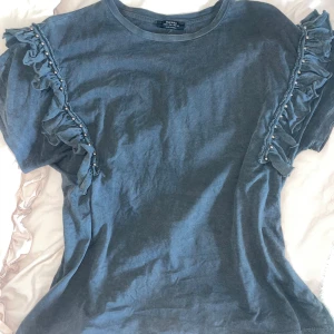 Grå t-shirt med volang och pärlor från Bershka - Säljer en blå t-shirt från Bershka med korta ärmar. Toppen har volangdetaljer längs axlarna och pärlor som dekoration. Köpte från bershka för 399 säljer för 150