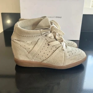 Beige mockasneakers med kilklack från Isabel Marant - Snygga beige sneakers i mocka från Isabel Marant med kilklack och snörning. Skorna har perforerade detaljer och en gummisula. Perfekta för dig som vill ha en sportig men ändå trendig look. Nypris 6000kr