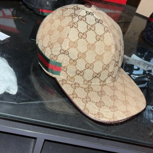 Beige keps från Gucci med GG-mönster - Säljer en beige keps från Gucci med klassiskt GG-mönster och grönt/rött band på sidan. Kepsen har böjd skärm och justerbar rem baktill. Perfekt för dig som gillar lyxiga accessoarer och vill sticka ut.