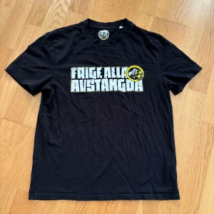 Svart AIK t-shirt med tryck 'Frige Alla Avstängda' - Svart AIK t-shirt med stort vitt och gult tryck på bröstet där det står 'Frige Alla Avstångda och en rund svartgul logga bredvid med handklor. Klassisk passform och rund halsringning. Perfekt för dig som gillar statement-plagg.