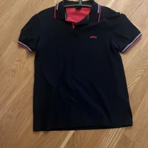 Mörkblå pikétröja från BOSS - Säljer en svart pikétröja från BOSS med röda och vita detaljer på krage och ärmslut. Tröjan har klassisk krage, korta ärmar och liten BOSS-logga broderad på bröstet. Perfekt för en stilren och sportig look.