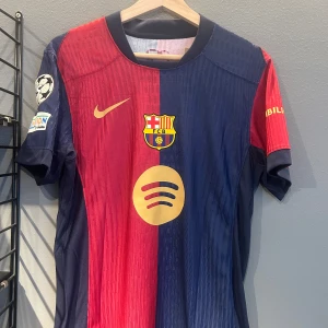 FC Barcelona matchtröja Nike Raphinha - Säljer en officiell FC Barcelona fotbollströja med Raphinha 11 på ryggen. Tröjan är röd och blå med guldiga detaljer, klubbmärke och sponsorlogga på bröstet. Kortärmad modell med Champions League-märke på ärmen. Helt ny med tagg en replika