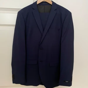 Mörkblå kostym - Säljer en stilren mörkblå kostym från Jack & Jones som varit använd enbart 2 gånger. Kavajen har två knappar framtill, klassiska fickor och slät finish. Kavajens storlek är 48 och byxorna är i storlek 46. Passar perfekt för killar i 15-17 års åldern. Säljer pga att den är för liten. Jag har även 2 st skjortor i storlek S från Jack & Jones som man kan få med för en liten extra slant. Pris kan diskuteras vid snabb affär.