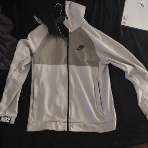 Grå hoodiejacka från Nike - Säljer en grå hoodiejacka från Nike med svart logga på bröstet och ärmslut. Jackan har dragkedja framtill, huva och två sidofickor. Perfekt för en sportig och avslappnad stil.