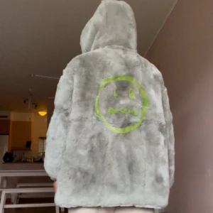 Grå pälsjacka med luva och smiley - Den här jackan är mycket varm och kan användas som en vinterjacka. Den har två sidor – ena sidan har fuskpäls som är mycket mjuk, och den andra sidan är vattentät. Du kan bära den på två olika sätt. Den väger 1,3 kg och är en mycket fin jacka.