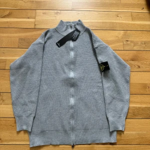 Grå stickad ziptröja från Stone Island - Säljer en grå stickad ziptröja från Stone Island med hel dragkedja och hög krage. Tröjan har lång ärm och klassisk Stone Island-logga på ena ärmen. Perfekt för lager-på-lager och stilren vardagslook.