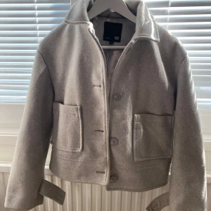 Ljusgrå kappa från 157 - Stilren ljusgrå kappa från 157 med klassisk krage och stora fickor framtill. Jackan är fodrad och har knäppning med knappar. Perfekt för dig som gillar en enkel och tidlös look.