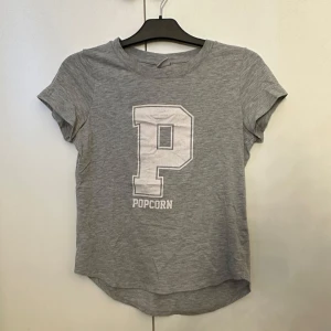 Grå t-shirt med tryck från Gina Tricot - Grå t-shirt från Gina Tricot med stort vitt P och texten 'Popcorn' på bröstet. Klassisk rund halsringning och korta ärmar. Tillverkad i mjuk ekologisk bomull för en skön känsla. Aldrig använd.