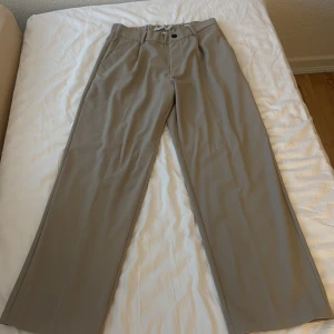 Beige kostymbyxor från Bershka - Snygga beige kostymbyxor från Bershka med raka ben och normal passform. Byxorna har hällor för bälte, knapp och dragkedja framtill samt en bakficka. Perfekta för en stilren look.