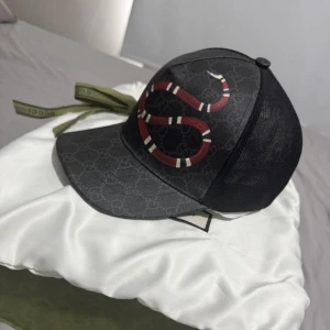 Svart keps från Gucci med ormmotiv - Snygg svart keps från Gucci med klassiskt GG-mönster och en röd-vit orm broderad framtill. Baksidan är i mesh för extra ventilation och har justerbart spänne med röd-gröna detaljer. Perfekt accessoar för en trendig look.
