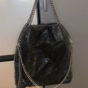 Svart axelväska med kedjedetaljer - Säljer en svart axelväska med glansig finish och silverfärgade kedjor längs kanterna och som axelrem. Väskan har en mjuk, slouchy form och är perfekt för att bära dina viktigaste saker. Snygg och trendig accessoar som lyfter varje outfit.