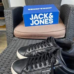 Svarta sneakers från Jack & Jones - Svarta sneakers från Jack & Jones med vita sulor och klassisk snörning. Liten reva på undersidan men det har två lager inuti så det är inget problem. Jag vill bara bli av med dessa skor. Priset kan diskuteras.