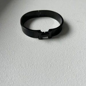 Svart armband med H-spänne - Stilrent svart armband med ett H-format spänne framtill. Armbandet har en blank finish och är tillverkat i metall, perfekt för att ge en modern touch till din outfit.