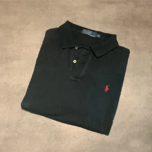 Svart pikétröja från Polo Ralph Lauren - Klassisk svart pikétröja från Polo Ralph Lauren med röd broderad logga på bröstet. Tröjan har korta ärmar, krage och knäppning med två knappar framtill. Perfekt för en stilren och avslappnad look. Tröjan är blekt på några ställen som du ser på bild 2 och 3. Därav lågt pris. Pris kan diskuteras! Bara att hörav av sig vid funderingar eller frågor!