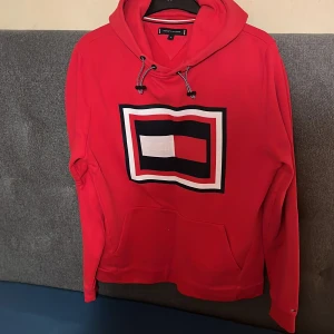 Röd hoodie från Tommy Hilfiger - Säljer en röd hoodie från Tommy Hilfiger med stor logga framtill i vitt, mörk blått och rött. Tröjan har huva med dragsko och en stor magficka. Perfekt för kyliga sommardagar den är i storlek M skulle säga 8,5av10 skick original pris ca 1800kr 