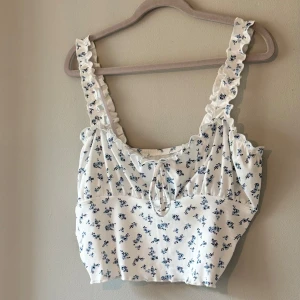 Vit blommig topp från Shein - Säljer en söt vit topp från Shein med blå blommor och volangdetaljer. Toppen har en knytning framtill och är ärmlös med smala axelband. Perfekt för en somrig look! 🌸