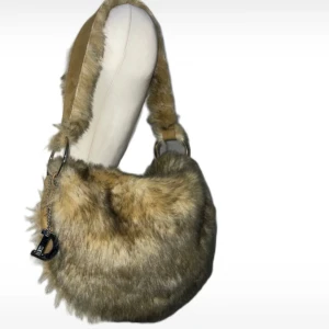 Beige fluffig axelväska med pälsimitation - Supermjuk och fluffig beige axelväska med pälsimitation och bred axelrem. Väskan har en silvrig D-detalj och rundad form, perfekt för att sticka ut. Insidan är klädd i mockaliknande material.