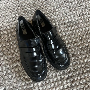 Svarta loafers från Zara - Säljer ett par klassiska svarta loafers från Zara i storlek 37. Skorna har en glansig finish och rund tå, perfekt för en stilren look. Passar till många olika outfits och är lätta att matcha.