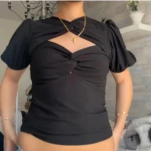 Svart topp med puffärmar  - Trendig svart topp med korta puffärmar och twistad detalj framtill som skapar en snygg öppning vid bröstet. Toppen har en figurnära passform och är perfekt för att lyfta en outfit.
