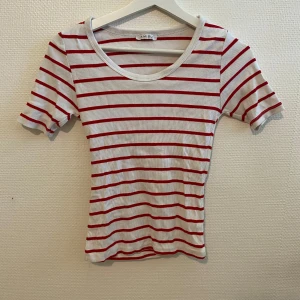 Randig T-shirt - Det står ingen storlek, jag skulle nog gissa på storlek XS. Säljer en vit ribbad T-shirt med röda horisontella ränder. T-shirten har rund halsringning och korta ärmar. Perfekt för en avslappnad stil.