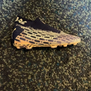 Puma Future fotbollsskor i gult och svart - Säljer ett par Puma Future fotbollsskor i gult och svart med coolt mönster och strumpliknande skaft. Skorna har snörlös design och är perfekta för dig som vill sticka ut på planen. Puma-logga på häl.