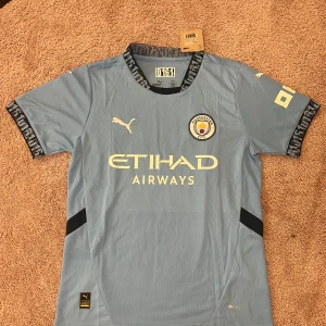 Manchester City hemmatröja Puma - Ljusblå Manchester City fotbollströja från Puma med klubbmärke och Etihad Airways-tryck på bröstet. Kortärmad modell med svarta detaljer och mönstrad krage. Buda gärna 