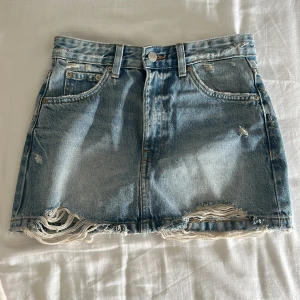 Blå jeanskjol från Pull&Bear - Säljer en snygg blå jeanskjol från Pull&Bear i storlek XS. Kjolen är kort med slitna detaljer och fransig nederkant för en trendig look. Klassisk femficksmodell med knapp och dragkedja framtill.