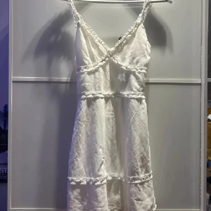 Vit omlottklänning med spetsdetaljer från H&M Divided - Söt vit kortklänning från H&M Divided med smala axelband och fina spetsdetaljer längs kanterna. Klänningen har omlottkänsla och volangdetaljer, perfekt för sommarens alla tillfällen.