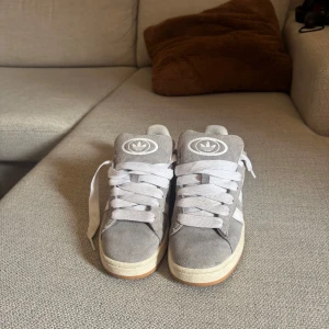Gråa sneakers från Adidas med vit sula - Säljer ett par gråa Adidas sneakers i mocka med vita detaljer och vit sula. Skorna har klassisk Adidas-logga på plösen och vita snören. Yttersulan är i gummi med mönster för bra grepp. Ny pris 1200kr 