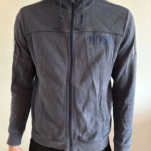 Blå zip-hoodie från Hugo Boss - Säljer en blå kofta från Hugo Boss med dragkedja och huva. Koftan har ett diskret rutigt mönster upptill. Broderad Hugo Boss-logga på bröstet. Perfekt för en avslappnad stil. 