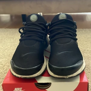 Nike Air Presto svart/vit - Säljer ett par svarta Nike Air Presto sneakers med vita detaljer och svart snörning. Skorna har en sportig design med rund tå och platt sula. Perfekta för dig som gillar stilrena och bekväma sneakers från Nike.