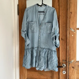 Ljusblå jeansklänning med volang från Zara - Supersöt ljusblå jeansklänning från Zara med volang nertill och knappar framtill. Klänningen har två bröstfickor och långa ärmar som kan rullas upp och fästas med knapp. Perfekt för en avslappnad och trendig look.