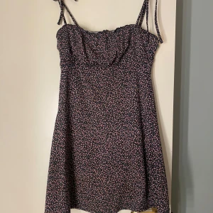 Blommig kortklänning med smala axelband - Supersöt kortklänning med smala knytbara axelband och blommigt mönster i rosa och vitt på mörkblå botten. Klänningen har en lätt utställd passform och är perfekt för varma dagar.